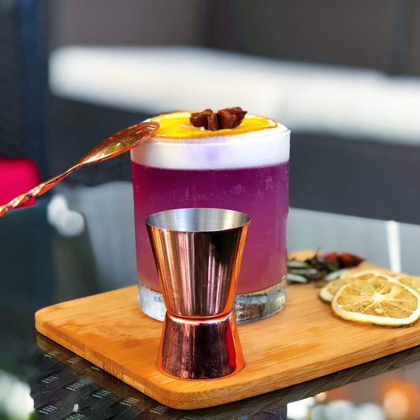 A vibrant purple cocktail with orange and star anise garnish, a copper jigger, and dried orange slices.