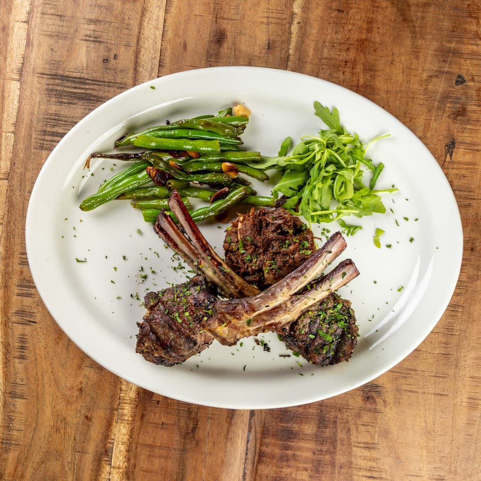 Grilled lamb chops with green beans, arugula, and shredded meat on a white plate.