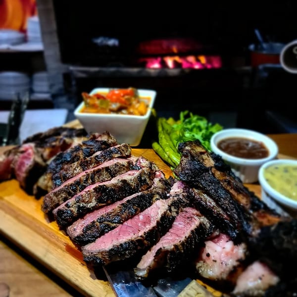 Slices of medium-rare grilled steak on a wooden board with sides and sauces, in front of a fireplace.