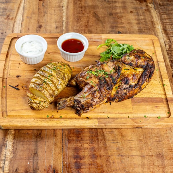 Grilled French chicken with a sliced baked potato, arugula, and two dipping sauces on a wooden board.