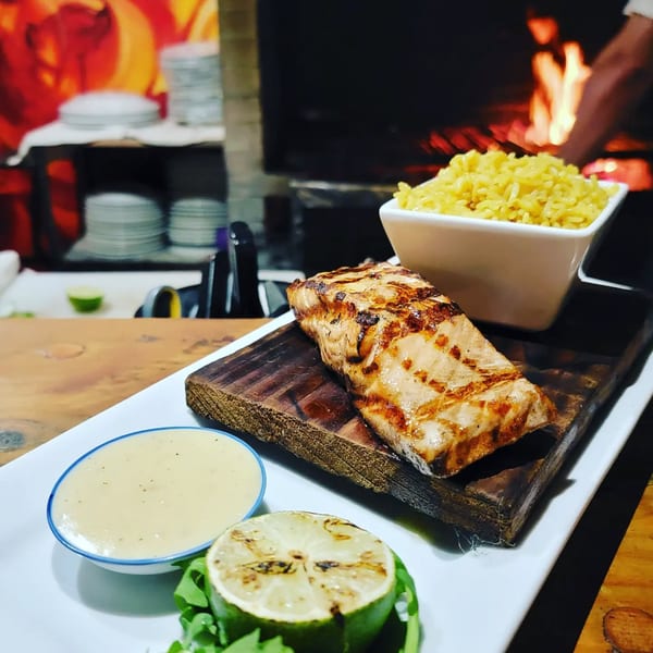 Grilled salmon on a wooden board with rice, sauce, and lime, in front of a fiery grill.